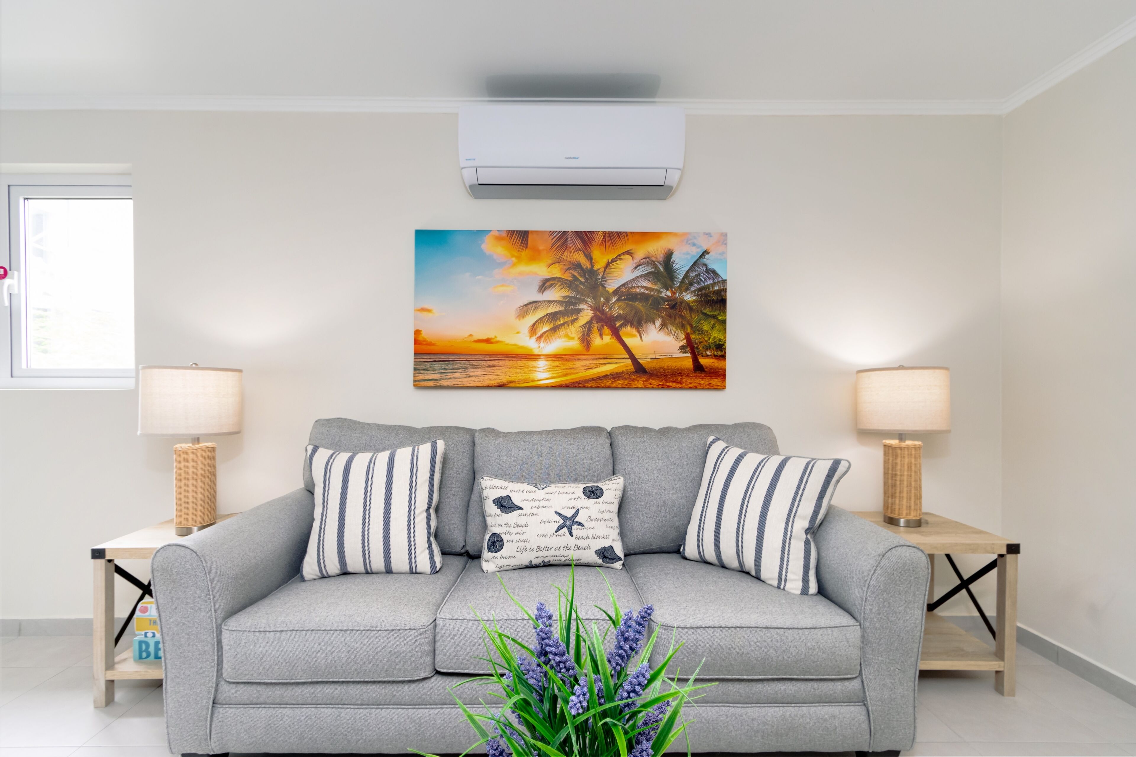 Een woonkamer met een grijze bank, gestreepte kussens, een wandairconditioner en een schilderij van een ondergaande zon met palmbomen.