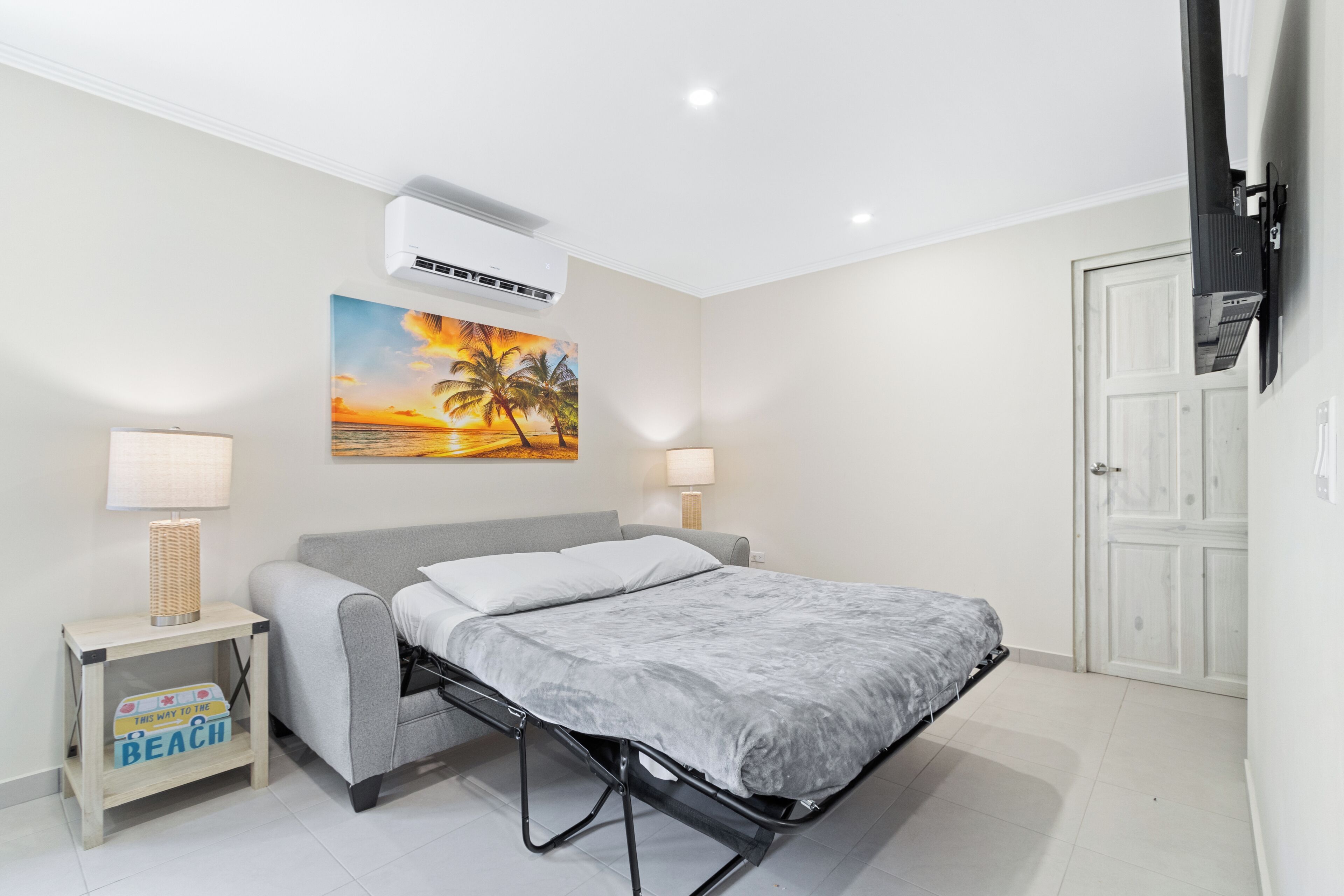 A bedroom with a bed, a nightstand, a lamp, a painting of a sunset, and a door.