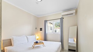 A hotel room with a large bed, a bedside table, a nightstand, a window with curtains, a mirror, and an air conditioning unit.
