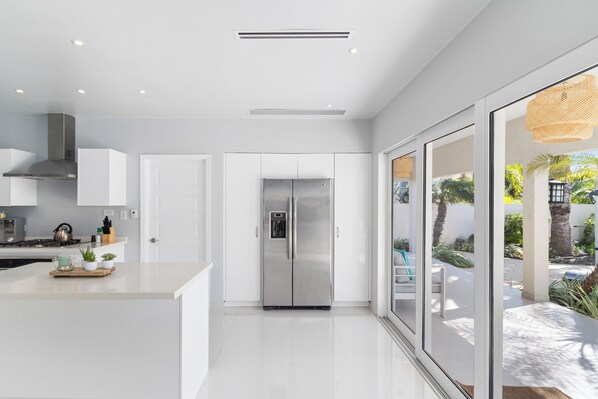 A modern kitchen with a stainless steel refrigerator, white cabinetry, and a large window overlooking a garden. - Opal 125 (Noord)
