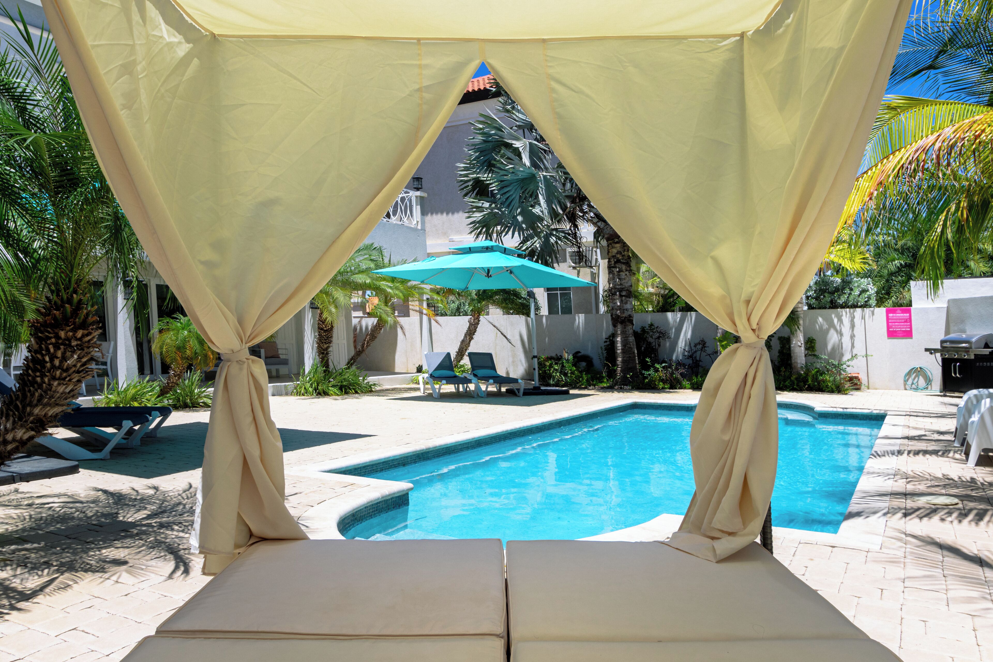A pool area with a canopy, lounge chairs, and a blue umbrella.
