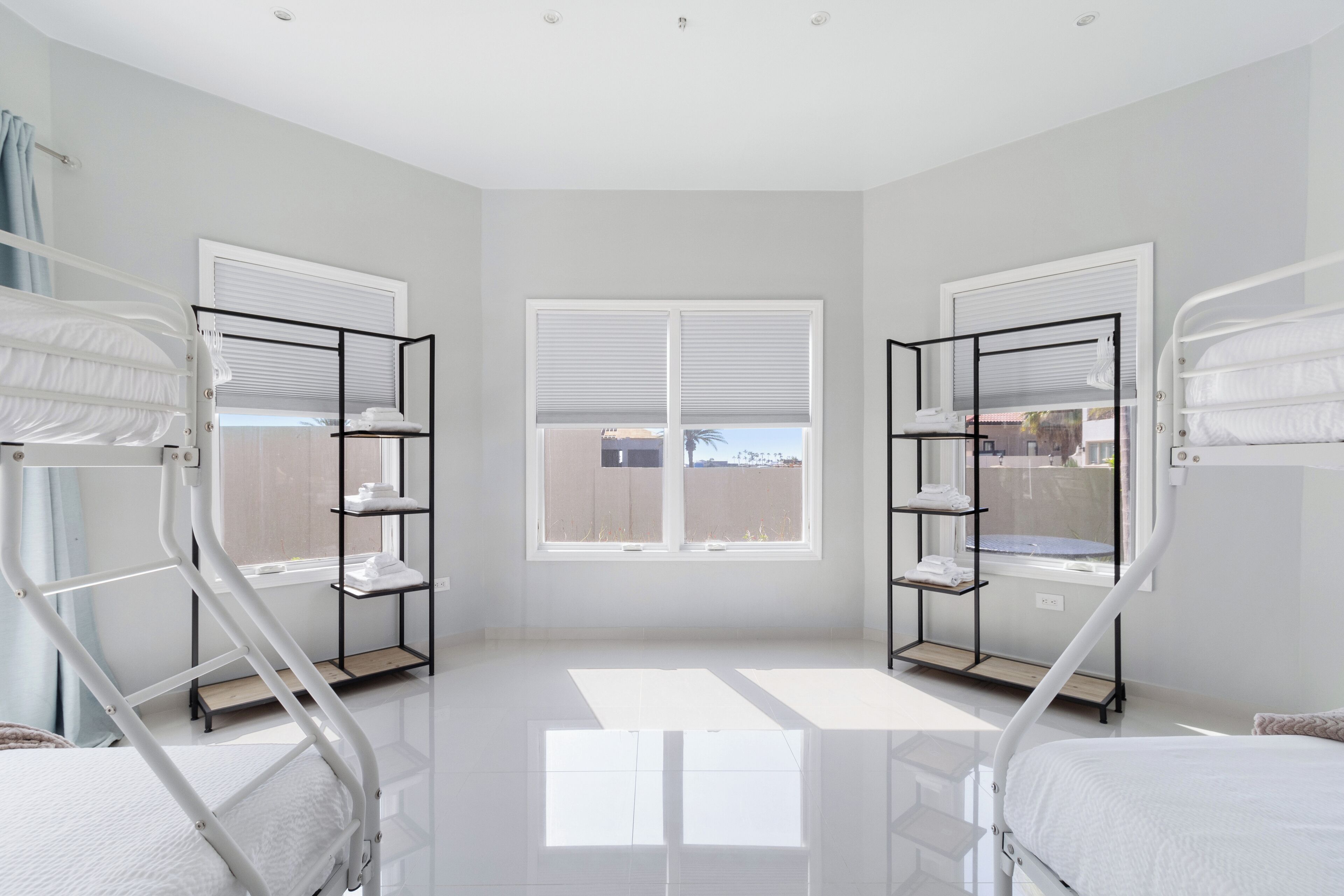 A bright, spacious room with two bunk beds, a window with blinds, and a shelf unit with towels.