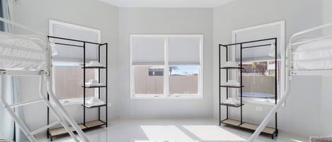 A bright, spacious room with two bunk beds, a window with blinds, and a shelf unit with towels.