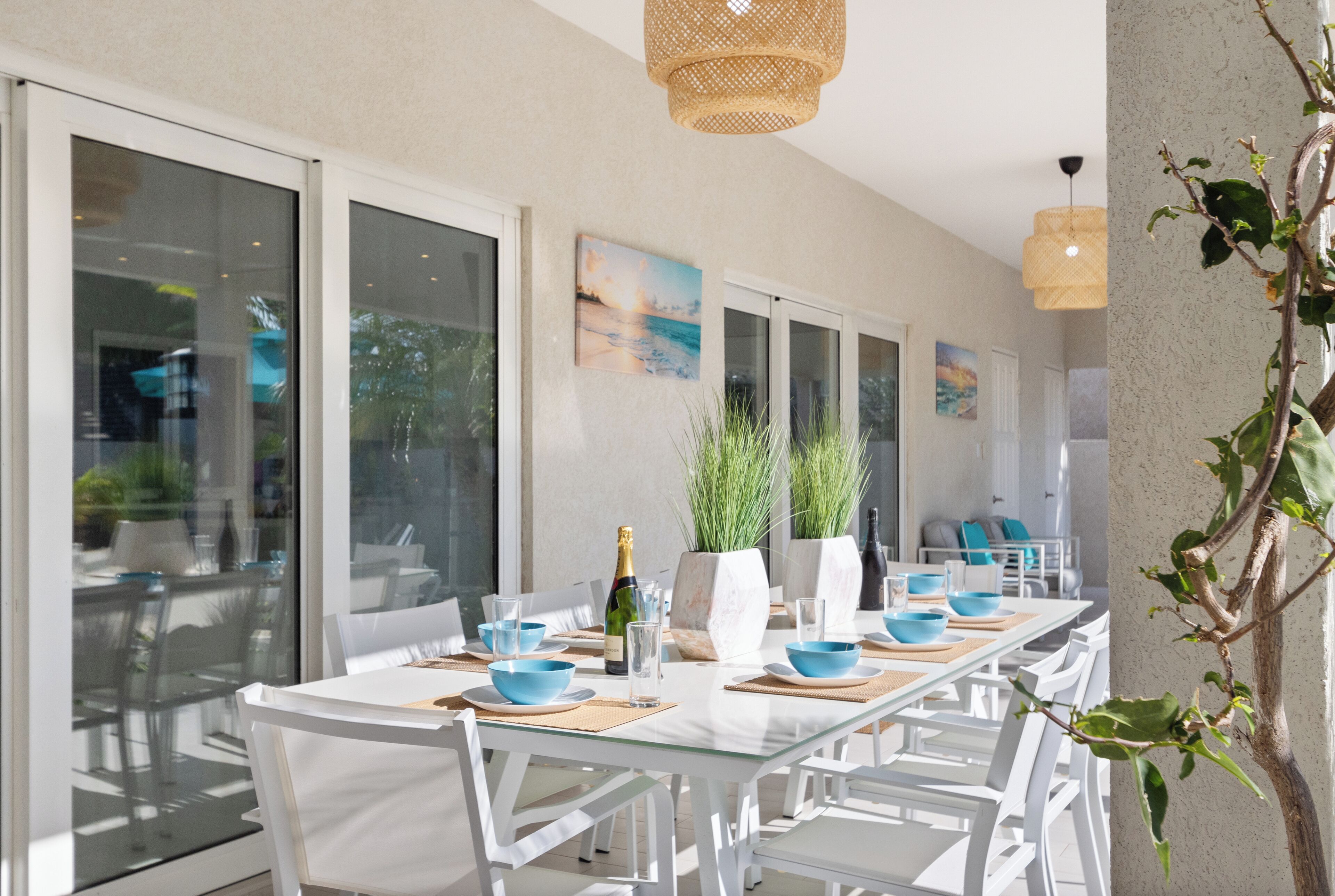 Un coin repas avec une longue table, des chaises blanches et des bols bleus. Des plantes en pot et une bouteille de champagne sont posées sur la table.