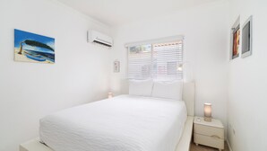 A bedroom with a bed, a nightstand, a wall-mounted air conditioner, and a window with blinds. - Little Paradise #5 - Barica Geel (Noord)