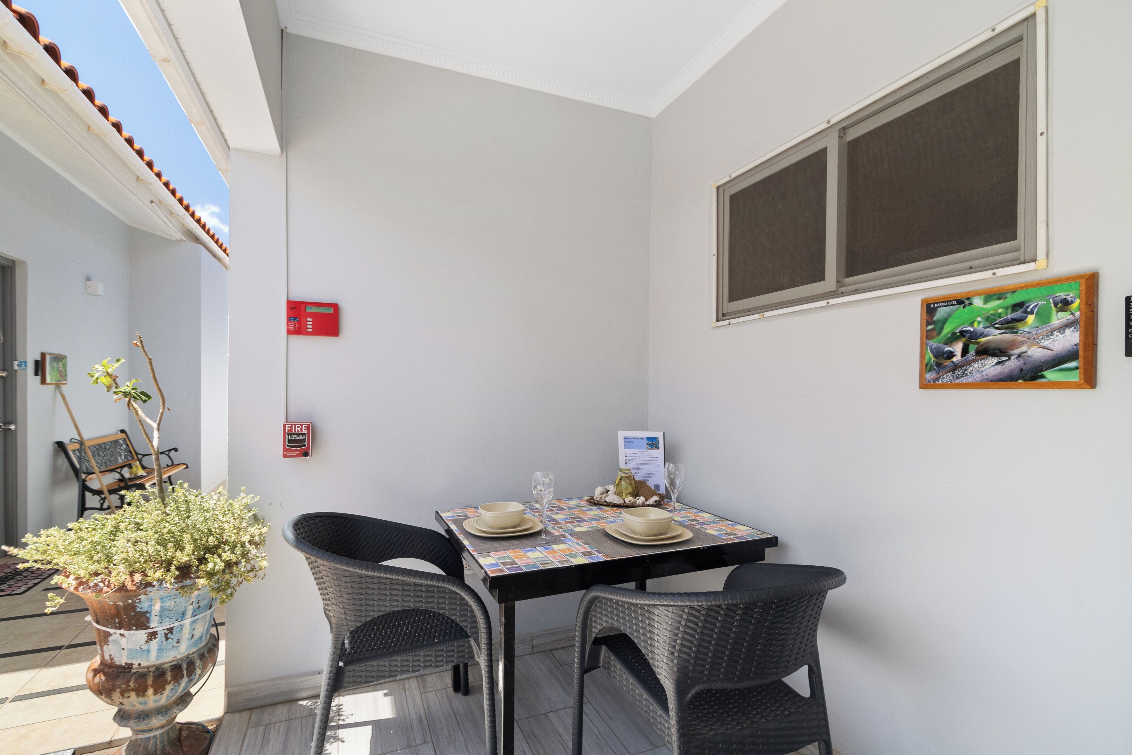 Un petit coin repas avec une table dressée pour deux personnes, deux chaises en osier, une plante en pot et un panneau indiquant l’emplacement du détecteur d’incendie sur le mur.