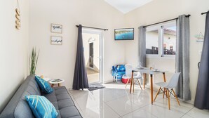 Ein modernes Wohnzimmer mit einem grauen Sofa, einem Esstisch mit Stühlen, einem kleinen Beistelltisch und Blick ins Freie durch eine Glastür.