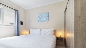 Ein Hotelzimmer mit einem großen Bett, zwei Nachttischen mit Lampen, einem Fenster mit Vorhängen und einem an der Wand hängenden Kunstwerk.