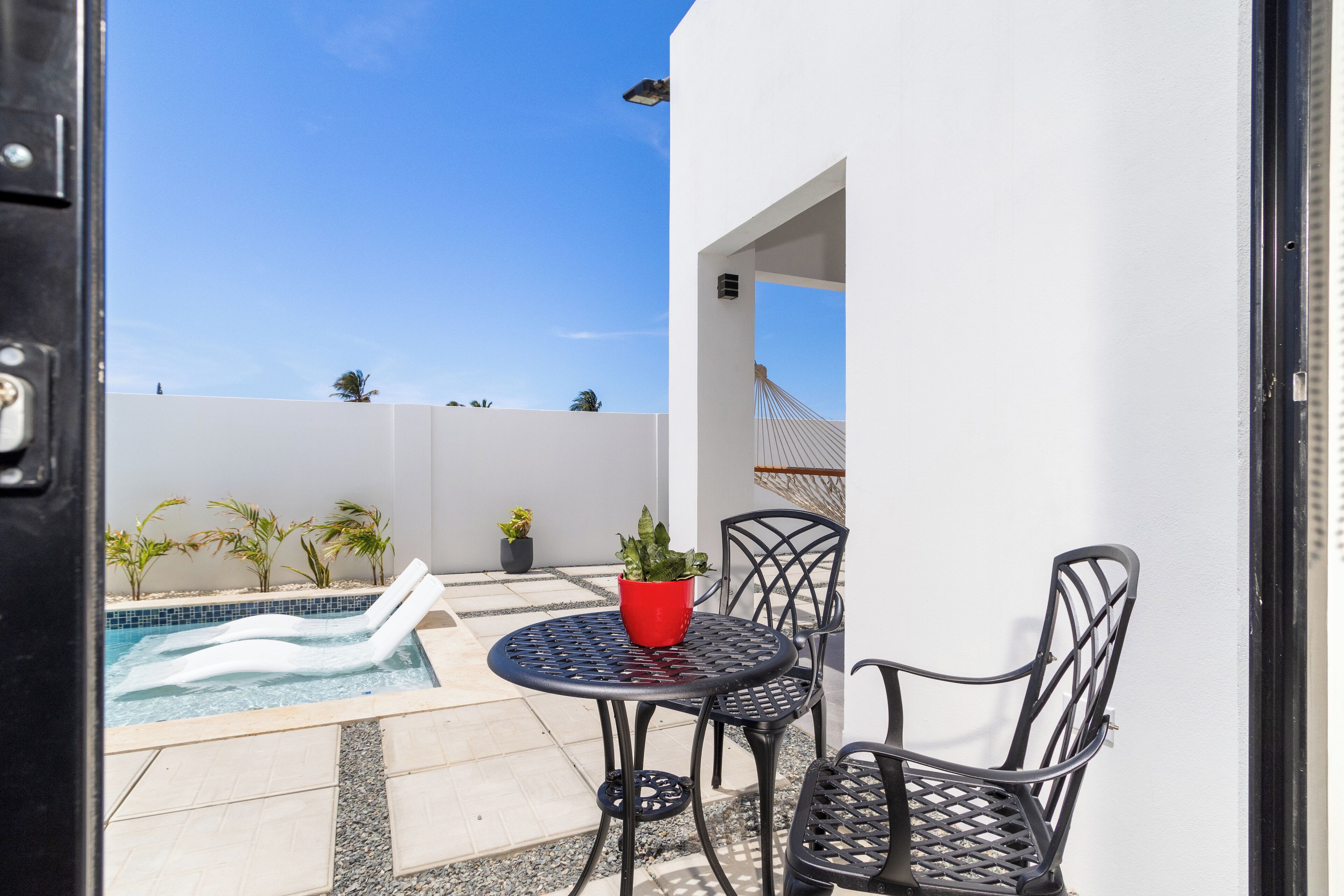 Un patio exterior con piscina, dos sillones de relax, una mesa con una maceta roja y sillas.