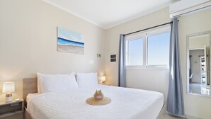 Uma cama bem arrumada com um chapéu decorativo, uma mesa de cabeceira com abajur, um quadro de uma praia e uma janela com cortinas.