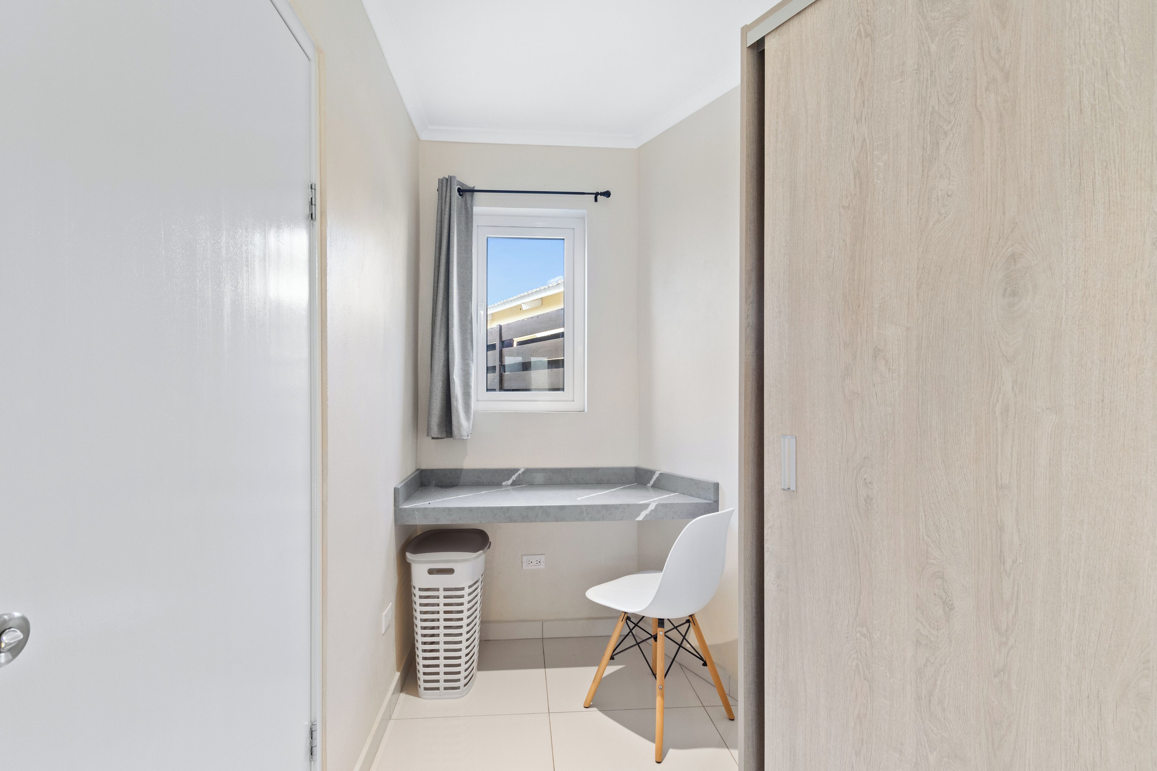 Une petite salle de bain moderne avec un lavabo blanc, une fenêtre avec un rideau, une chaise blanche et une poubelle.