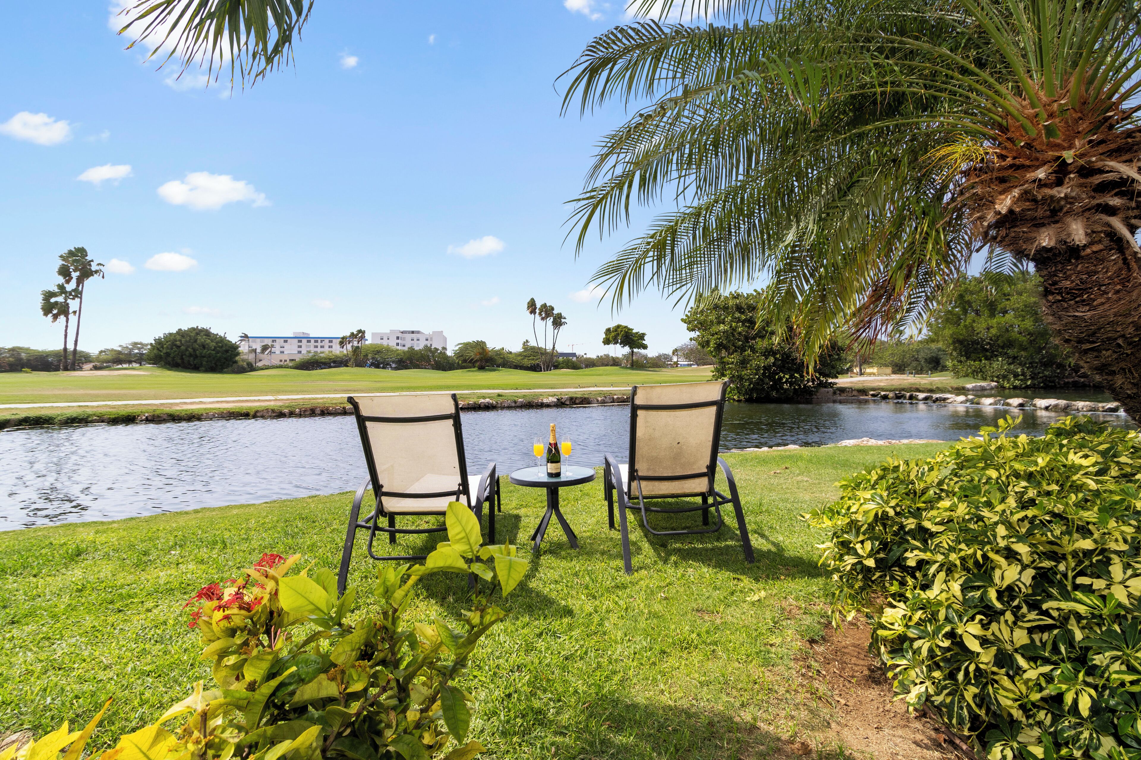 Ein ruhiger Außenbereich zum Sitzen mit zwei Stühlen und einem Tisch, mit Blick auf ruhiges Gewässer und einen Golfplatz im Hintergrund.