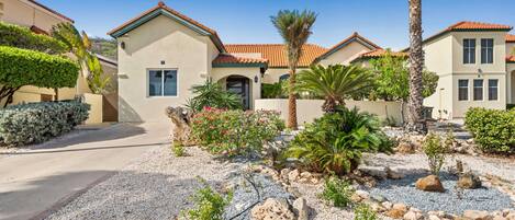 A residential area with houses featuring Mediterranean-style architecture, a gravel driveway, and landscaped gardens with various plants and rocks.
