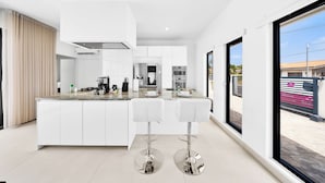 A modern kitchen with a central island, white cabinetry, stainless steel appliances, and a large window offering a view of the outside. - Koyari 35-f (Noord)