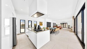 A modern kitchen with a central island, large windows, and a dining area with chairs and a table. - Koyari 35-f (Noord)