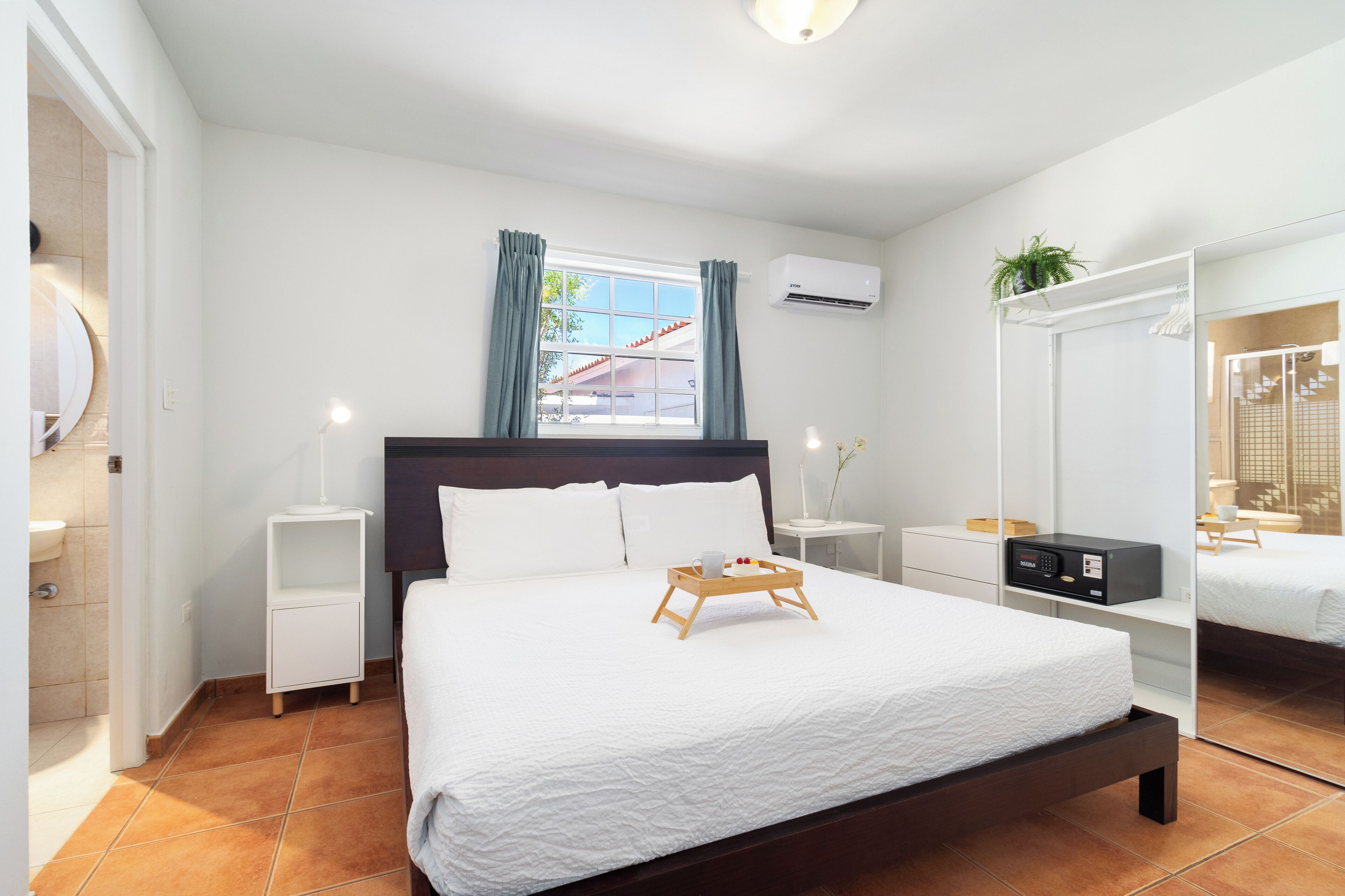A modern bedroom with a large bed, a bedside table with a lamp, a window with curtains, an air conditioning unit, and a wardrobe with a mirror.