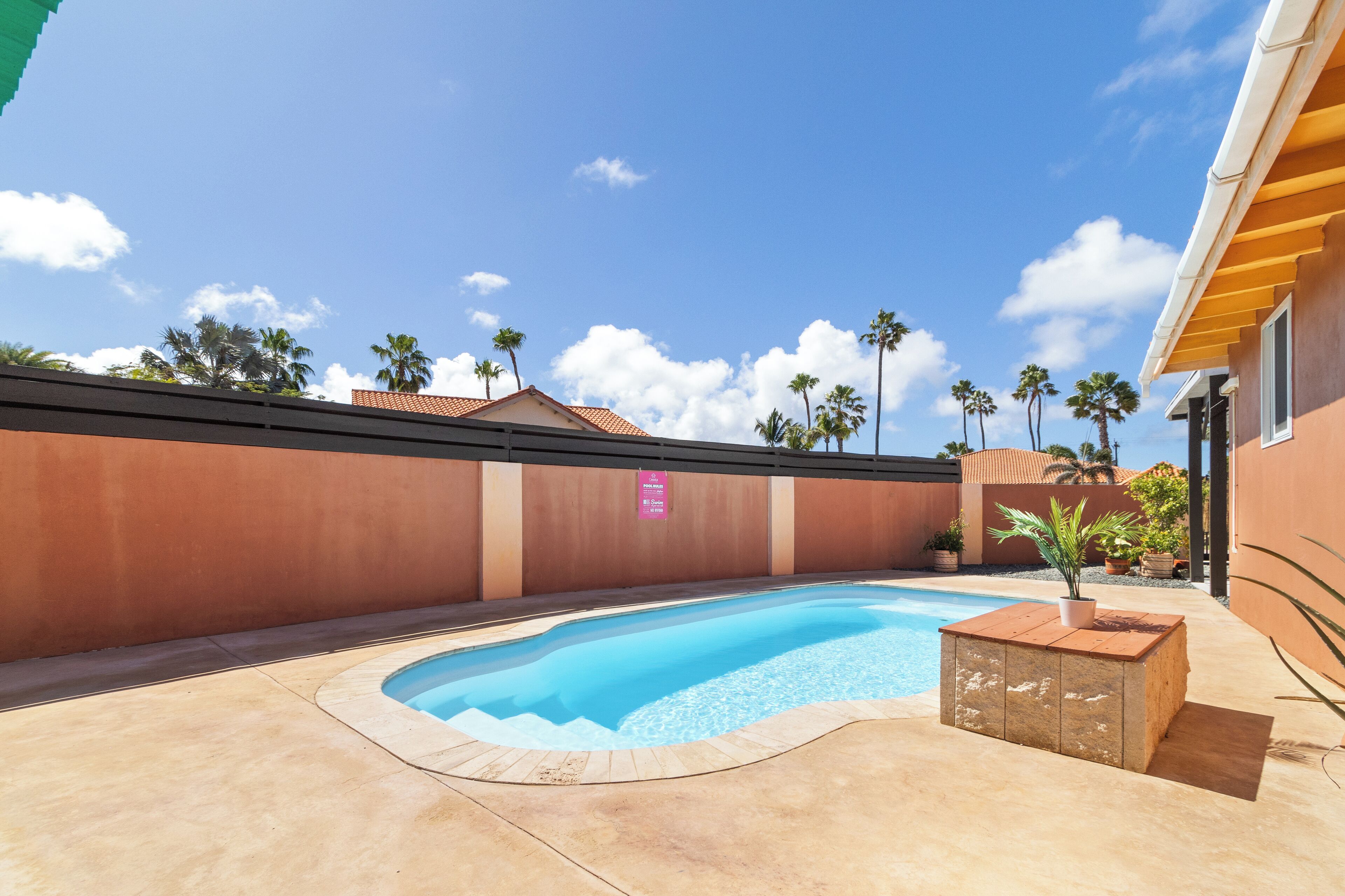 Une piscine dotée d’une terrasse en bois, entourée d’un petit muret et de palmiers.