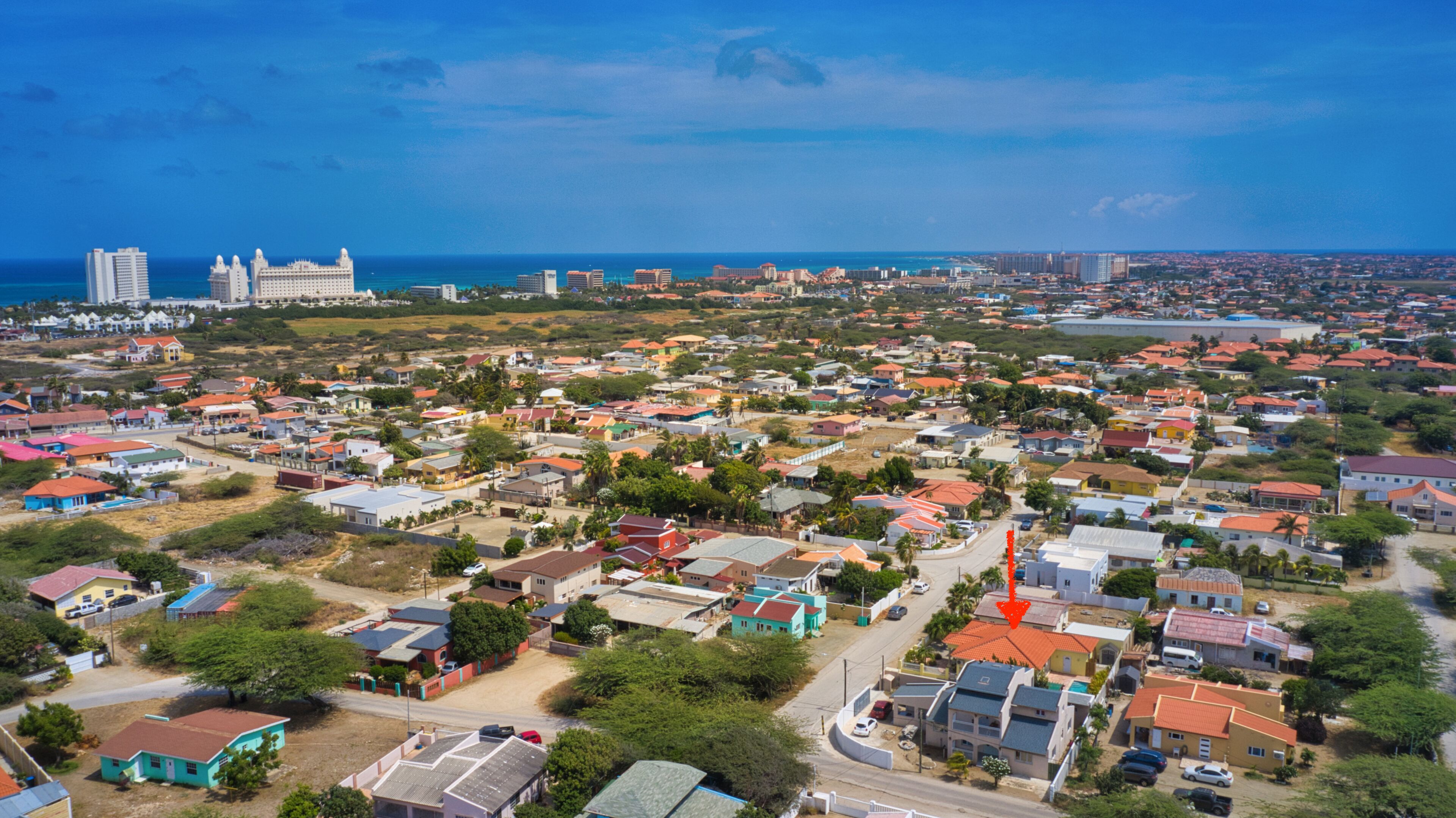 An aerial view of a coastal town with a mix of residential and commercial buildings, a prominent hotel with a large white facade, and a clear view of the ocean.