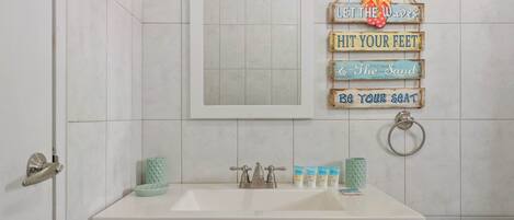 Une salle de bain avec un lavabo blanc, un miroir et des toilettes. Un panneau est fixé au mur, sur lequel on peut lire les inscriptions « Laissez la vague vous emporter » et « Sentez la puissance de la vague ».
