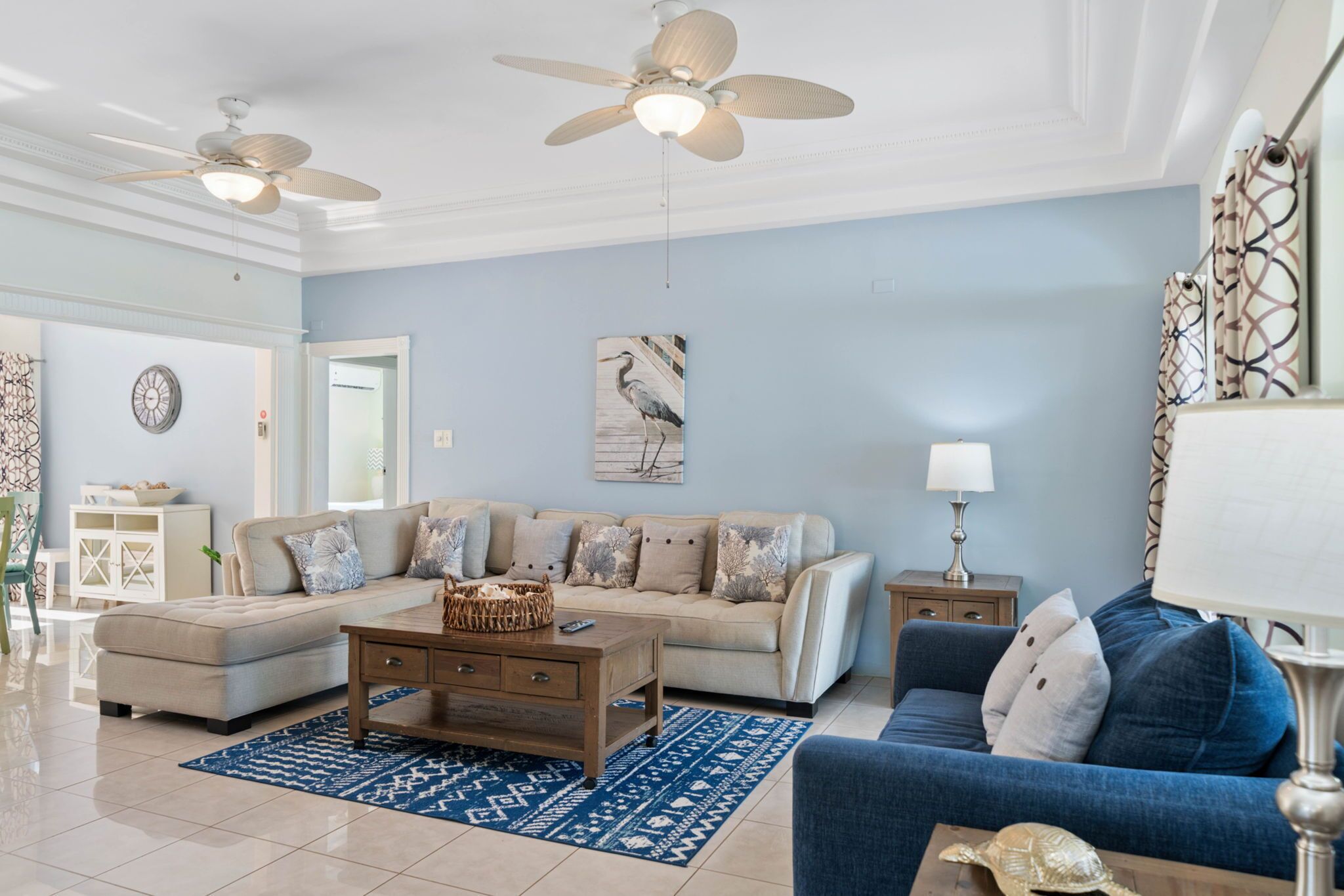 A spacious living room with a beige sectional sofa, a blue armchair, and a wooden coffee table. There are two ceiling fans, a wall-mounted lamp, and a framed artwork above the sofa.