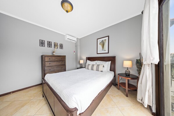 A bedroom with a bed, a nightstand, a dresser, a wall-mounted air conditioner, and a window with curtains.