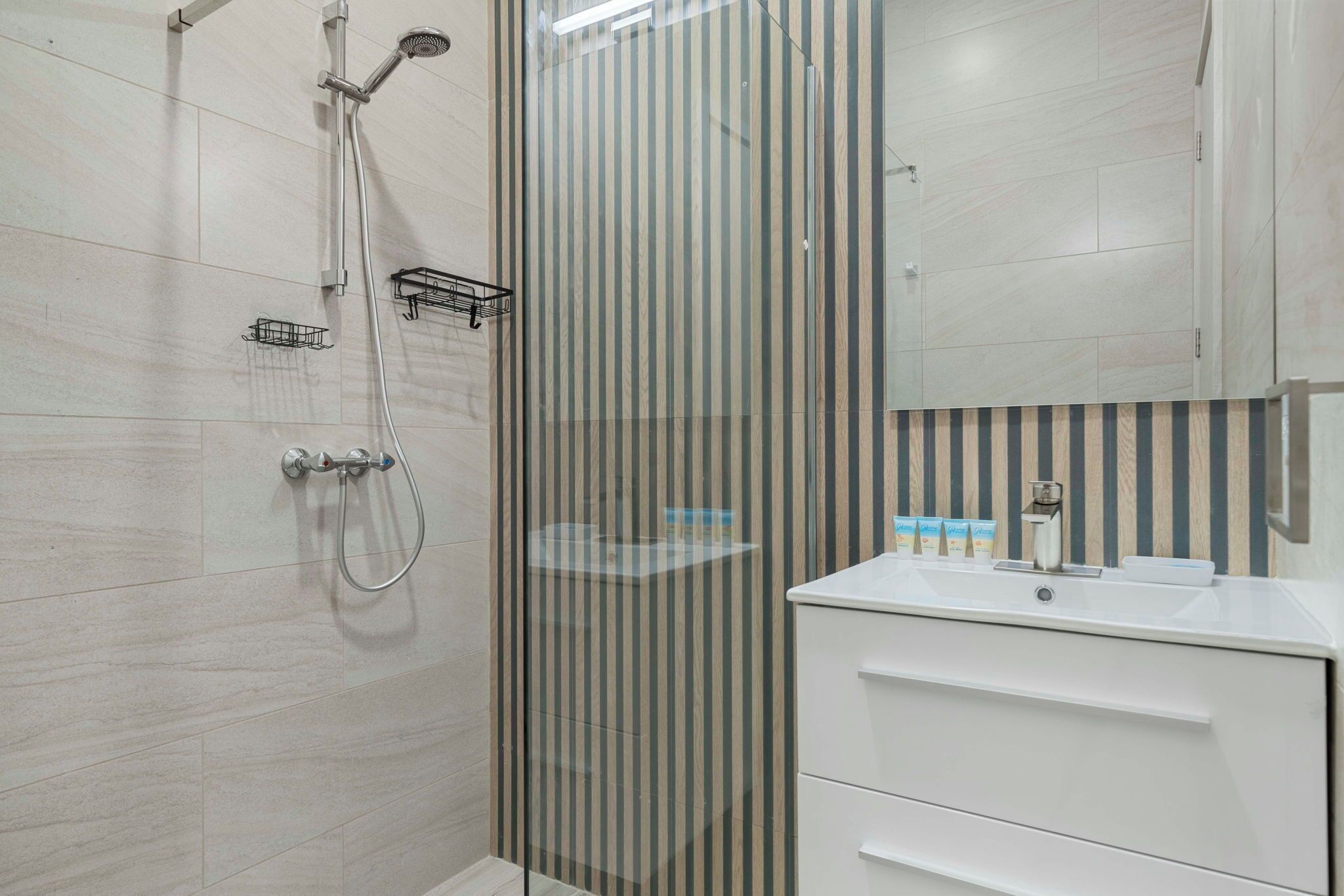 Een moderne badkamer met een douchecabine van glas, een witte wastafel met spiegel en gestreepte wandtegels.