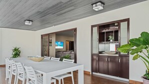 Een eethoek met een lange tafel en witte stoelen, een houten plafond en een donker houten kast met een plant.