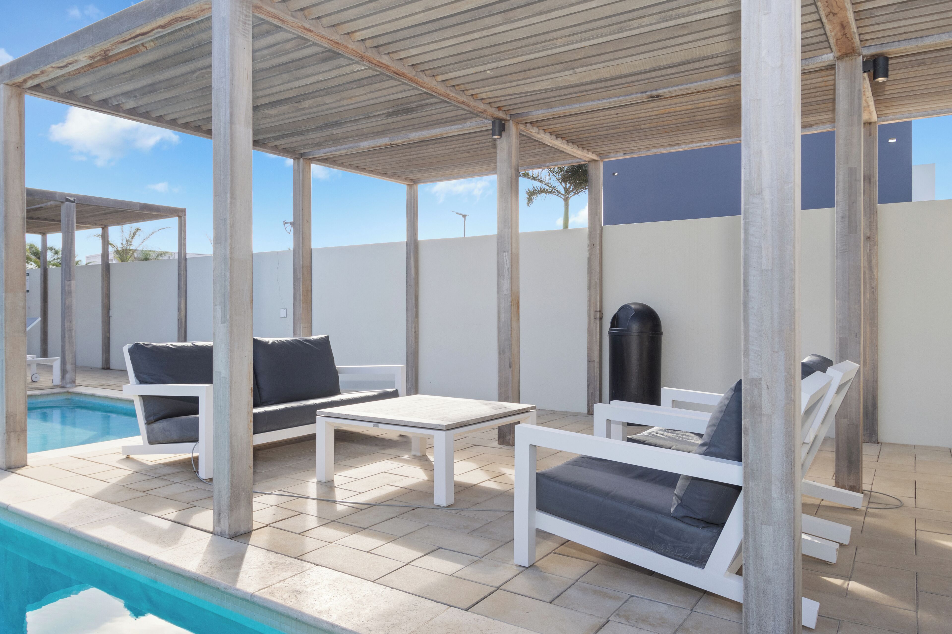 A covered outdoor seating area with a pool, white furniture, and a pergola structure.