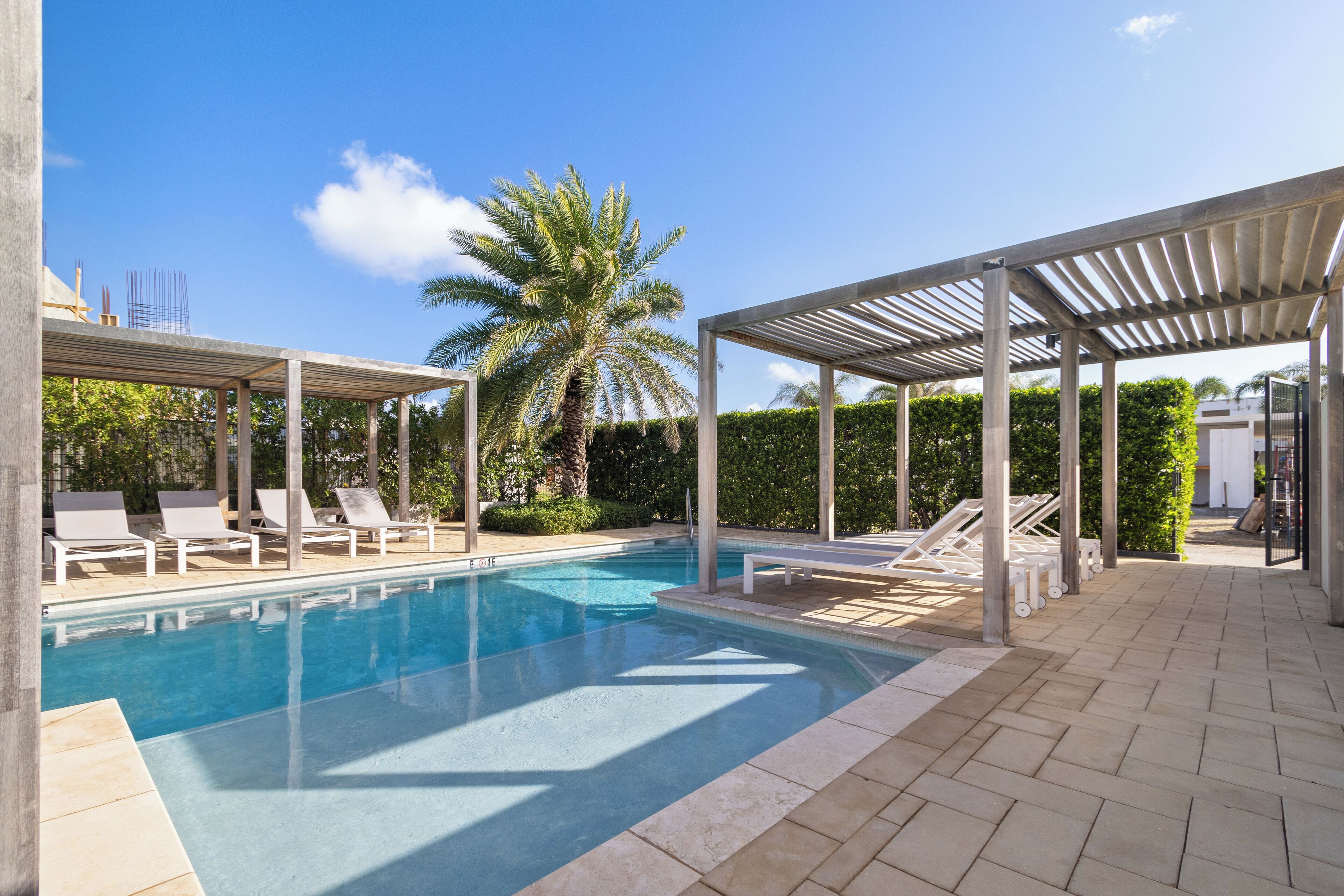 A swimming pool with lounge chairs and a covered area, surrounded by greenery and a clear sky.