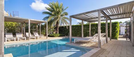 A swimming pool with lounge chairs and a covered area, surrounded by greenery and a clear sky.