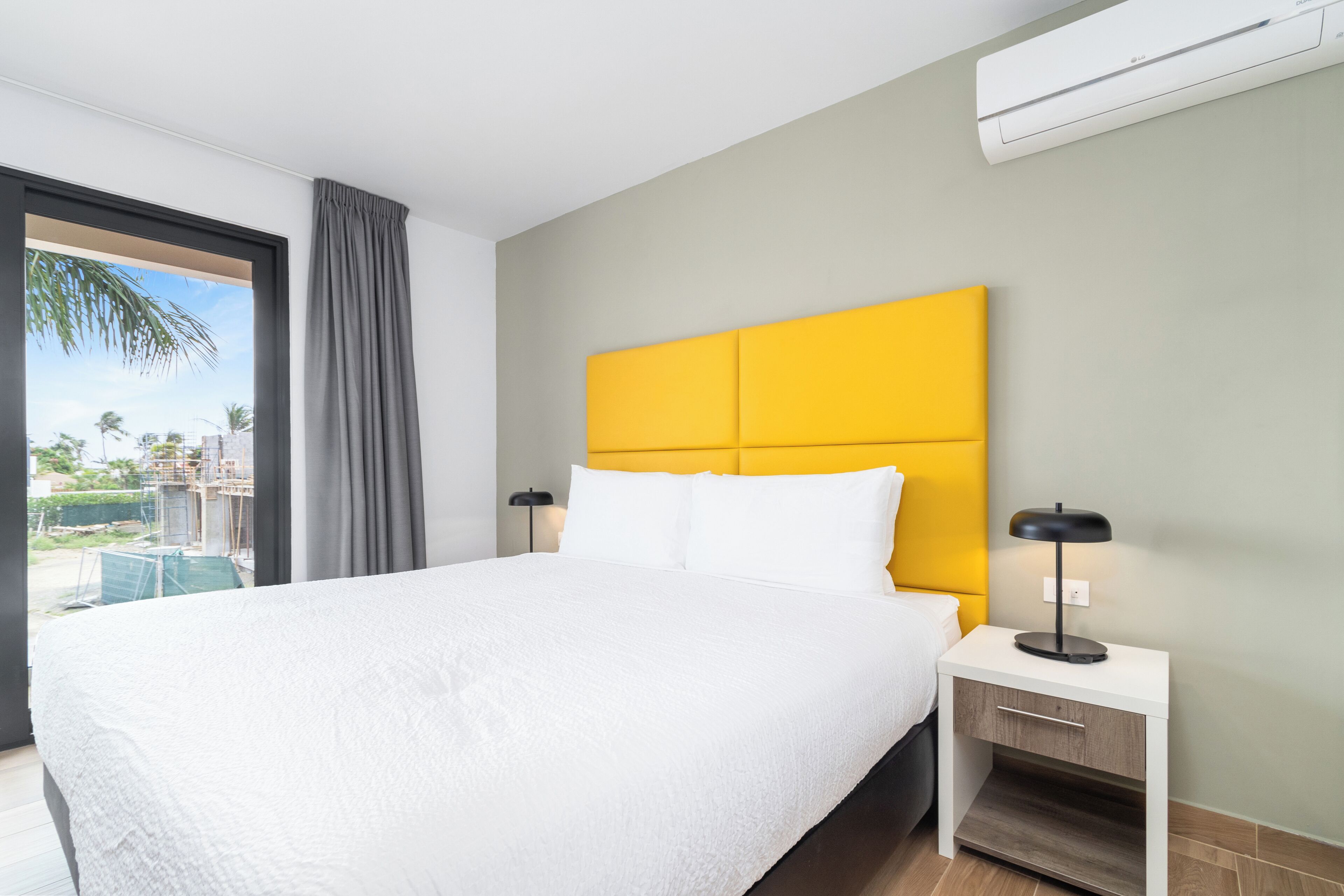 A modern hotel room with a large bed, a nightstand with a lamp, and a view of palm trees and buildings through a large window.