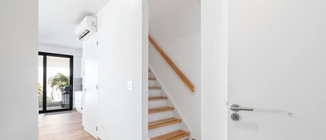 Un interior modern amb una escala de fusta, parets blanques i una porta de vidre que dona a un pati.