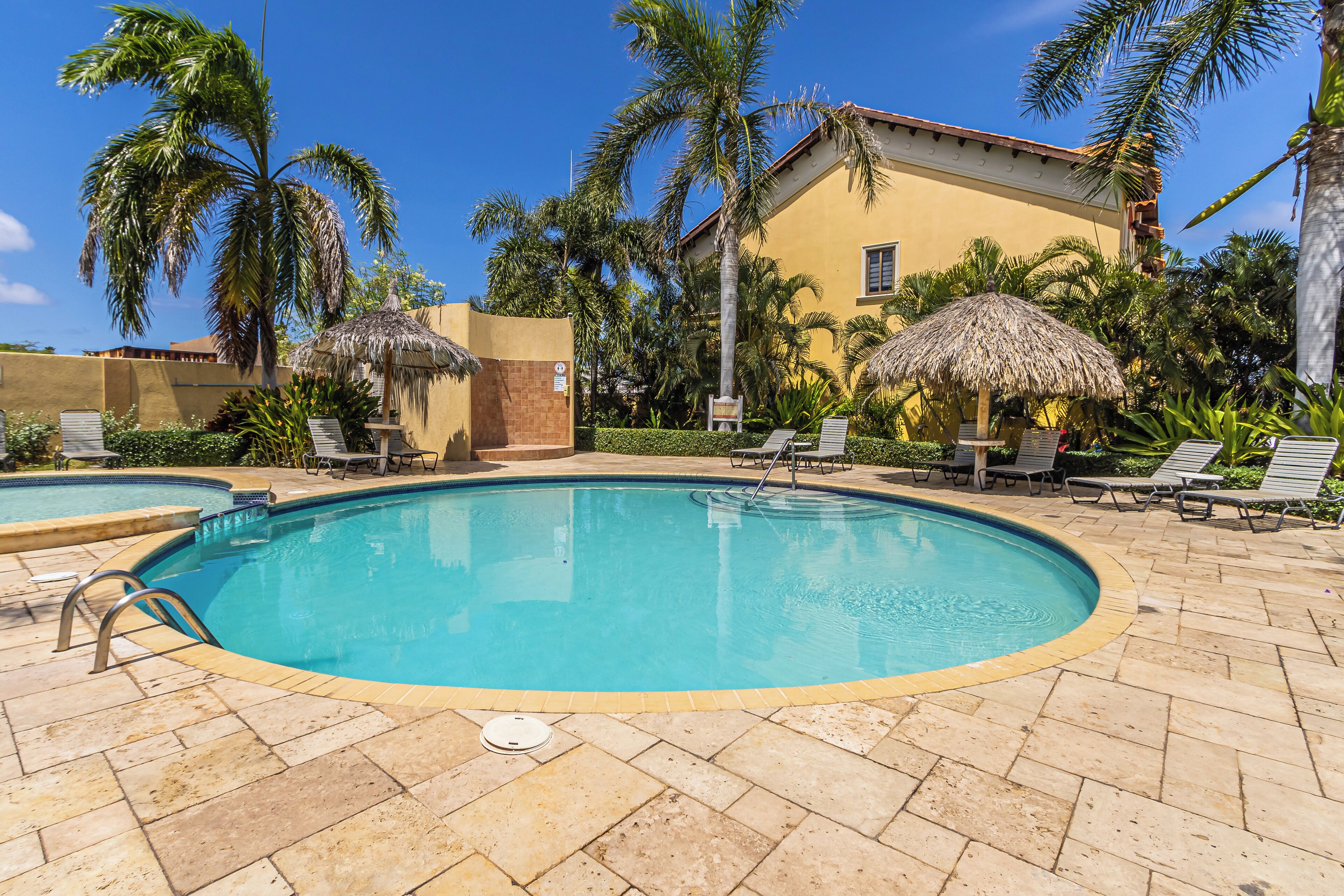A swimming pool with lounge chairs and thatched umbrellas, surrounded by palm trees and a building.