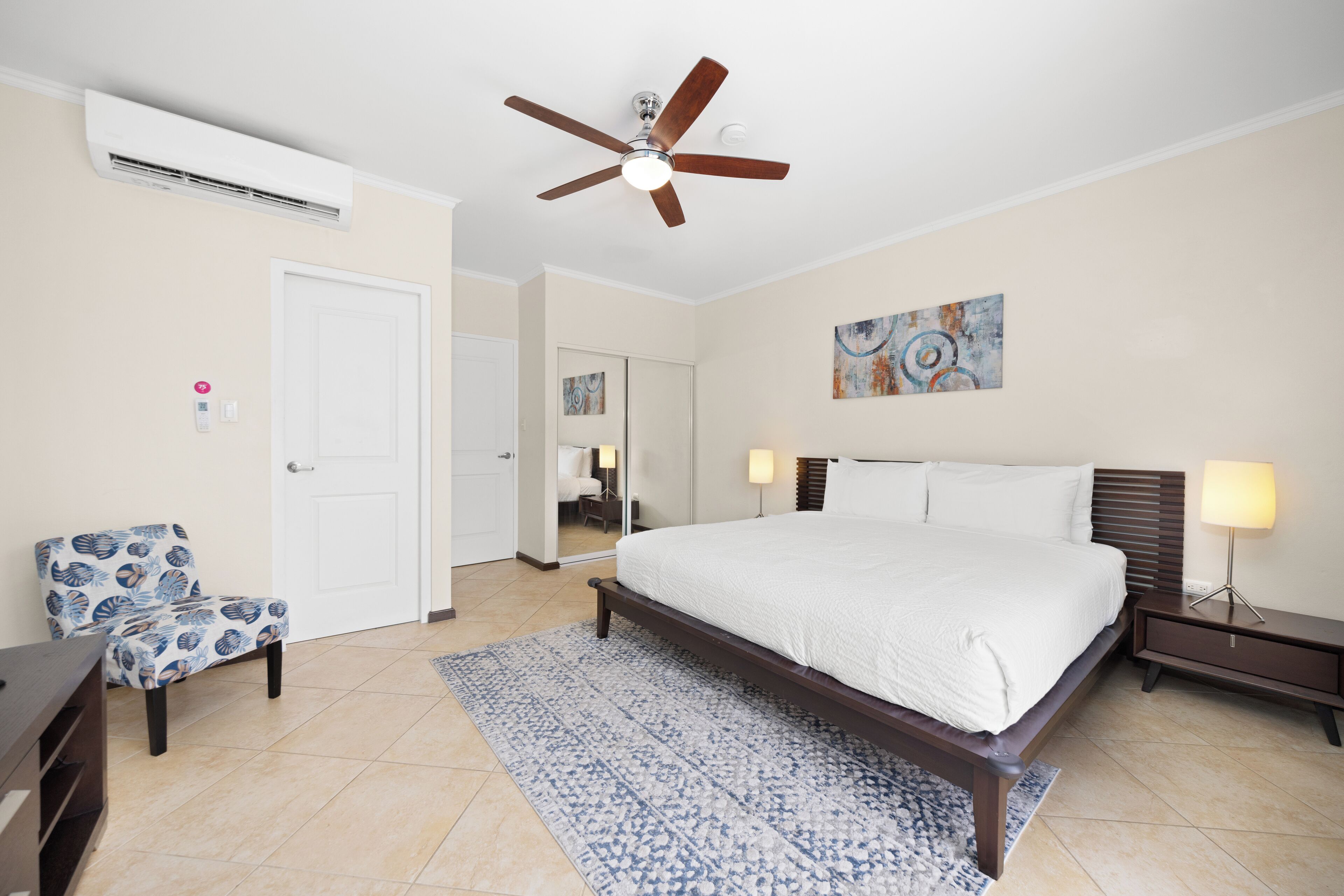 Un dormitorio con cama, mesitas de noche, ventilador de techo, unidad de aire acondicionado, una silla y un cuadro en la pared.