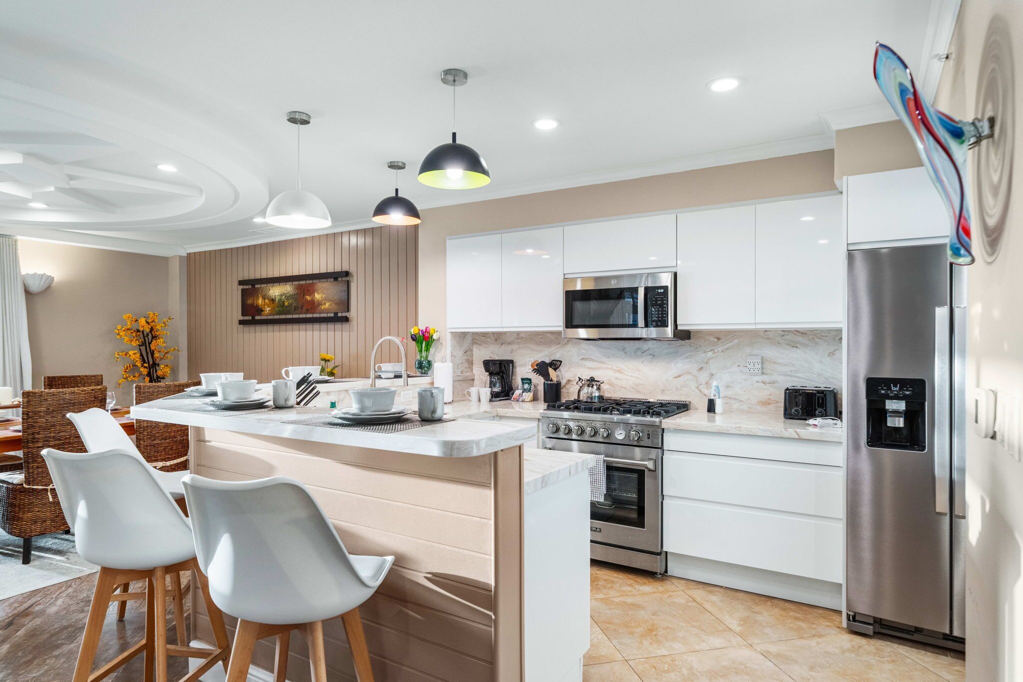 Una cucina moderna con mobili bianchi, elettrodomestici in acciaio inossidabile, un'isola centrale con sgabelli da bar, illuminazione a sospensione e un'area pranzo con mobili in vimini.