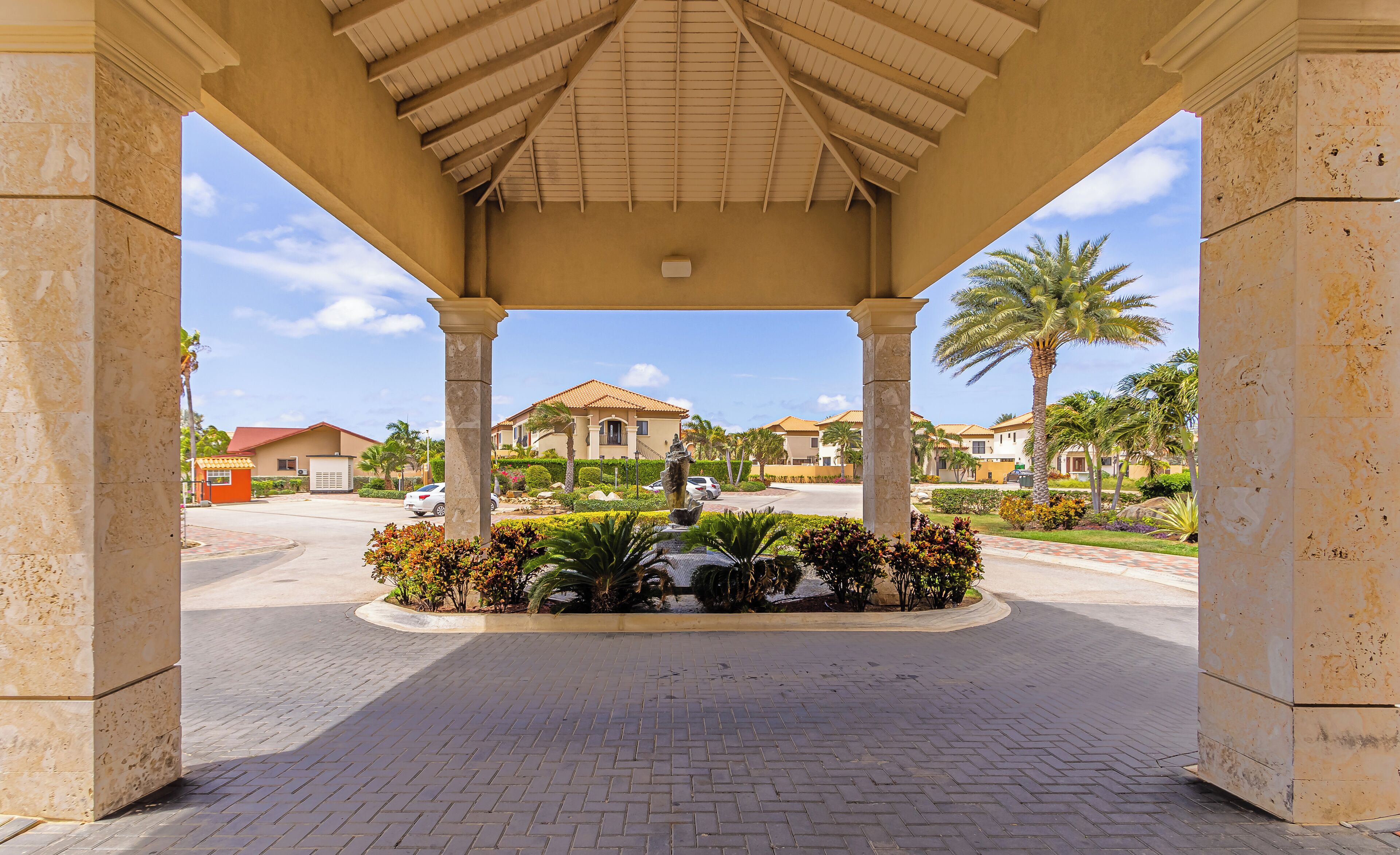 A covered entrance with a paved pathway leading to a residential area with houses and palm trees.