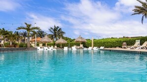 Une piscine bordée de chaises longues et ombragée par des parasols en feuilles de palmier, le tout entouré de palmiers et sous un ciel dégagé.