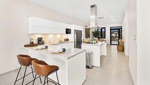 A modern kitchen with white cabinetry, a central island with bar stools, stainless steel appliances, and a large window providing natural light. - Catalina Residence Apt 3 - L.g. Smith Boulevard 546-j (Noord)