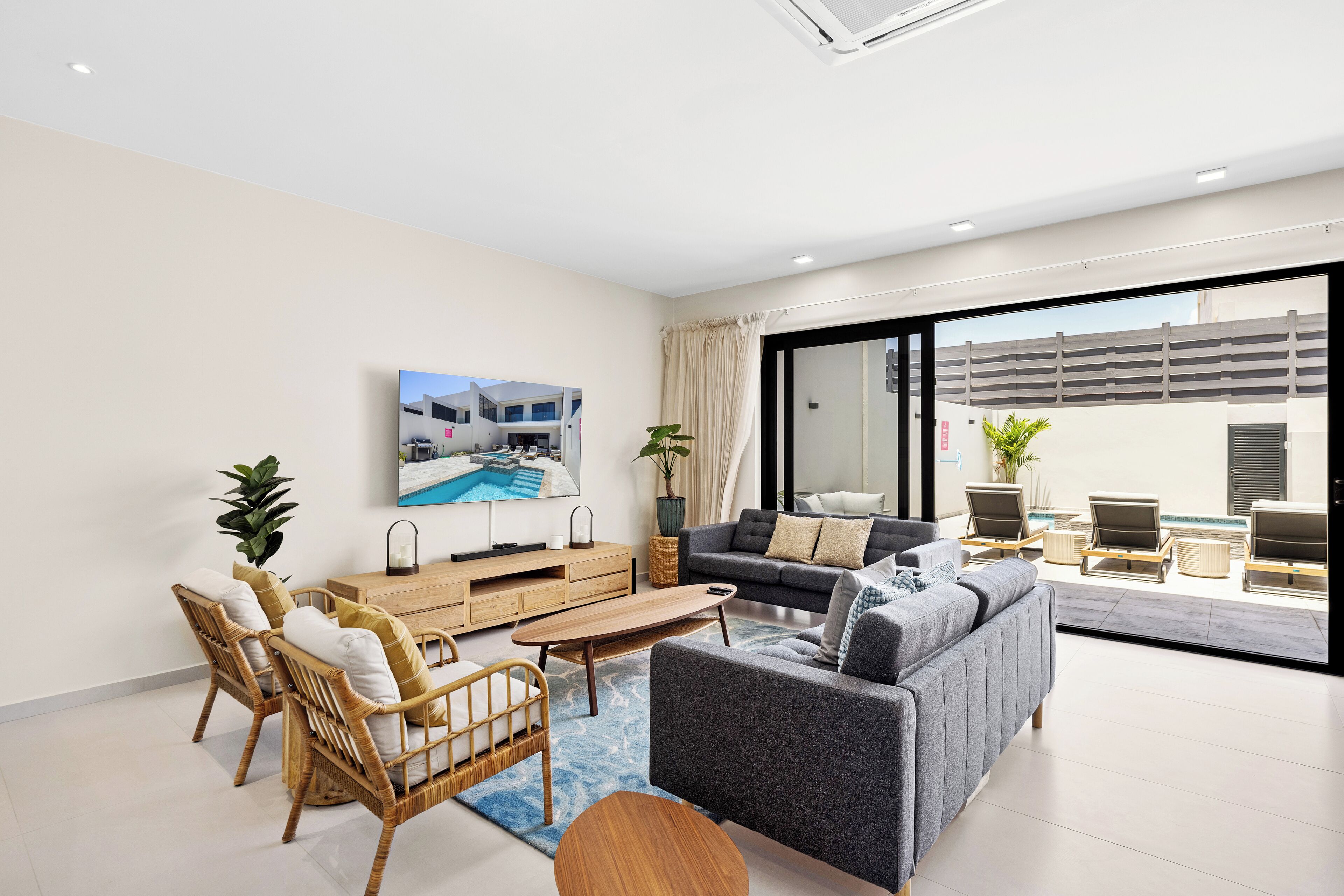 Een moderne woonkamer met een bank, fauteuils, een salontafel en een televisie. Er is een schuifdeur die leidt naar een buitenruimte met stoelen en een zwembad.