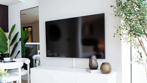 A modern living room with a large flat-screen TV mounted on the wall, a white console table with decorative items, and a dining area with a white table and chairs. - StayShort - Sky Gardens Studio with Skyscraper Views in DIFC! (DIFC)