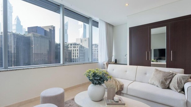 A modern living room with a large window, a white sofa, a round coffee table, and a dark wooden wardrobe. - StayShort - Sky Gardens Studio with Skyscraper Views in DIFC! (DIFC)