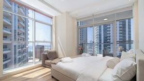 Quarto moderno com uma cama grande, um sofá e vista para edifícios altos através de janelas do chão ao teto.