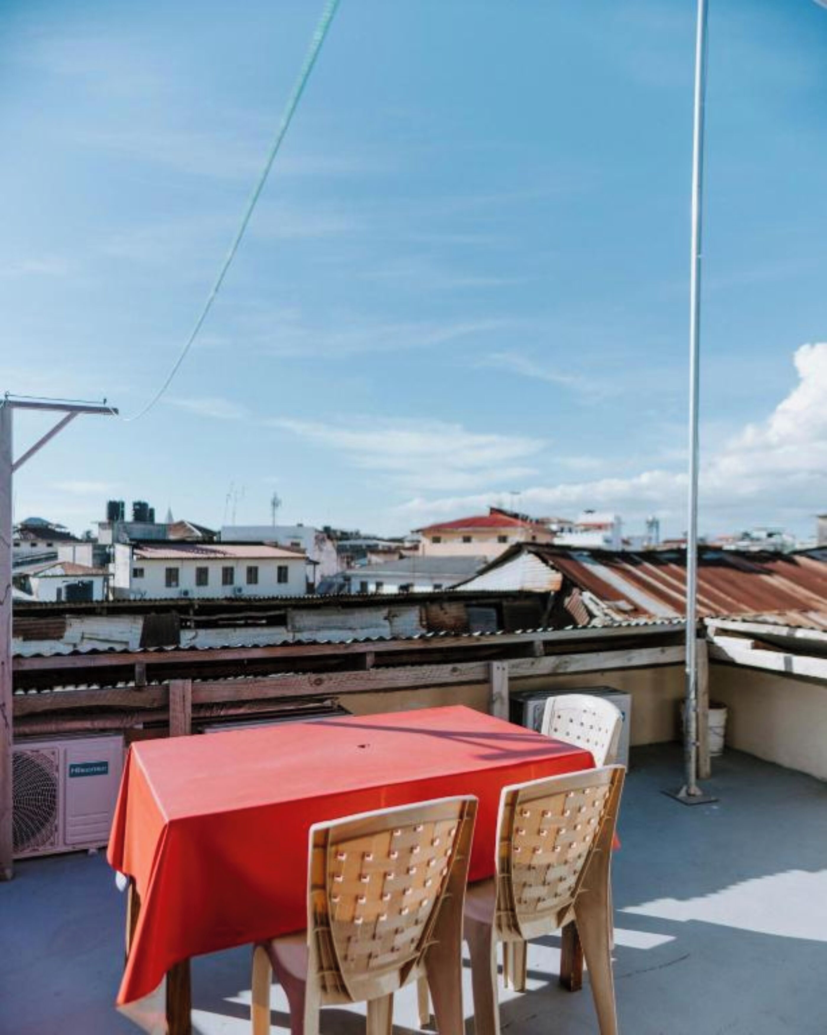 Terraza en la azotea con una mesa roja y dos sillas de mimbre, con vistas a un paisaje urbano con varios edificios.
