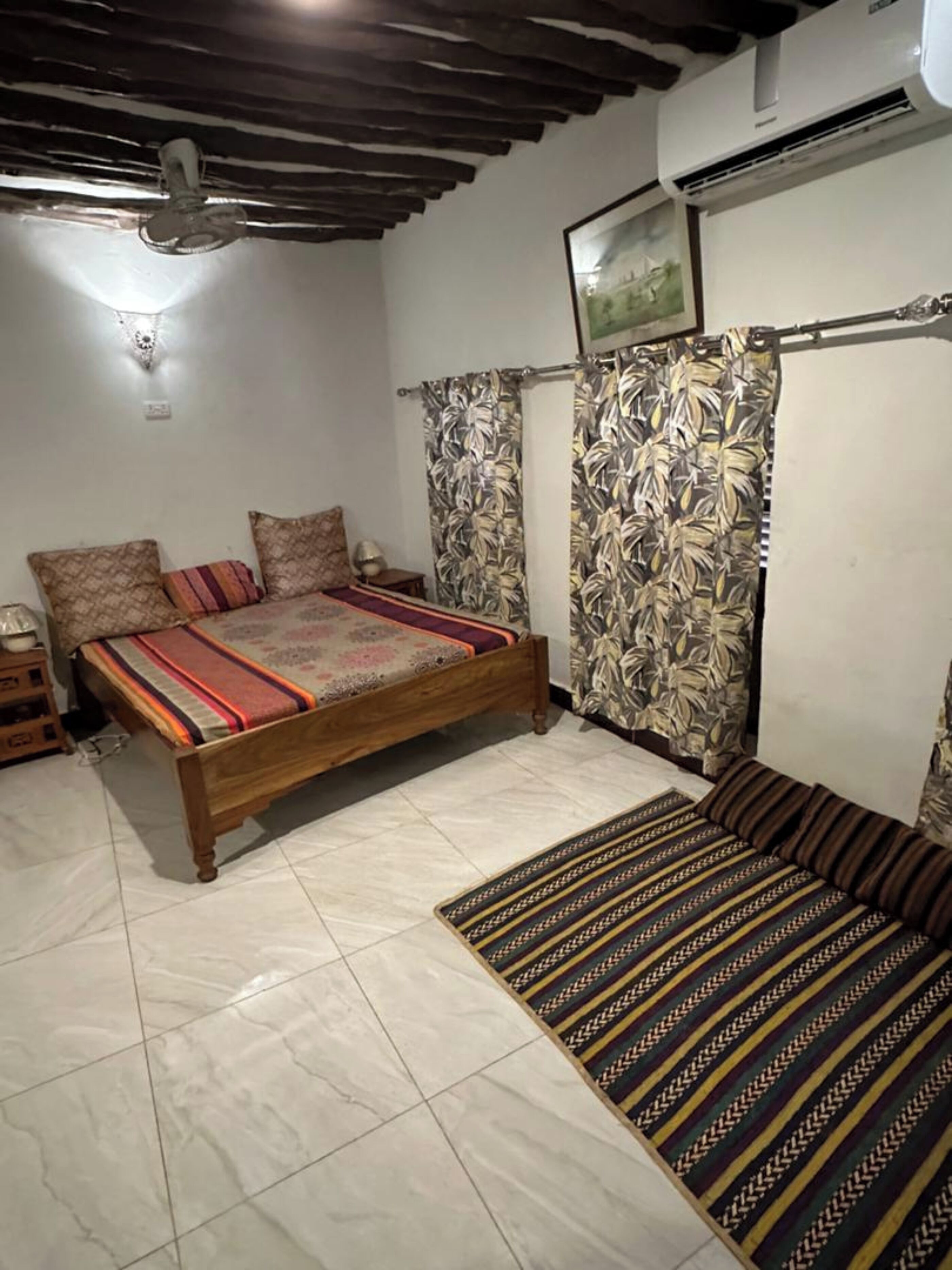 Ein Schlafzimmer mit einem hölzernen Bett, gemusterten Vorhängen, einem Fliesenboden und einem Deckenventilator.