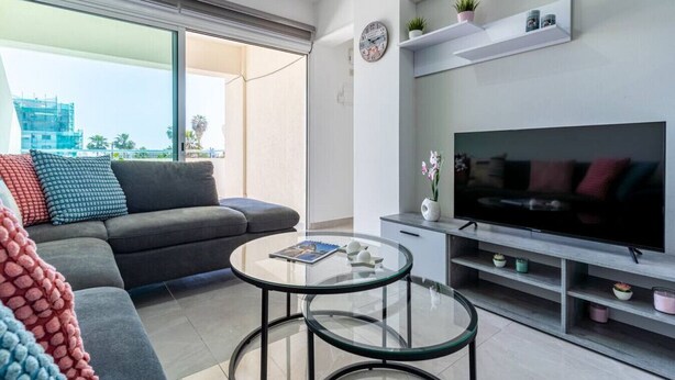 A modern living room with a grey sofa, a glass coffee table, a flat-screen TV, and a balcony with a view of the city. - Andrea 3-Bedroom Apt in Mackenzie W/ Pool (Cyprus - Larnaca)