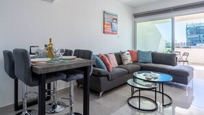 A modern living room with a dining area, a gray sofa, a glass coffee table, and a large window with a view of the city. - Andrea 3-Bedroom Apt in Mackenzie W/ Pool (Cyprus - Larnaca)