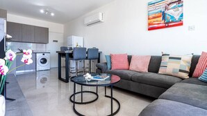 A modern living room with a grey sofa, a round glass coffee table, and a dining area with bar stools. There is a kitchen in the background with a washing machine and a dining table. - Andrea 3-Bedroom Apt in Mackenzie W/ Pool (Cyprus - Larnaca)