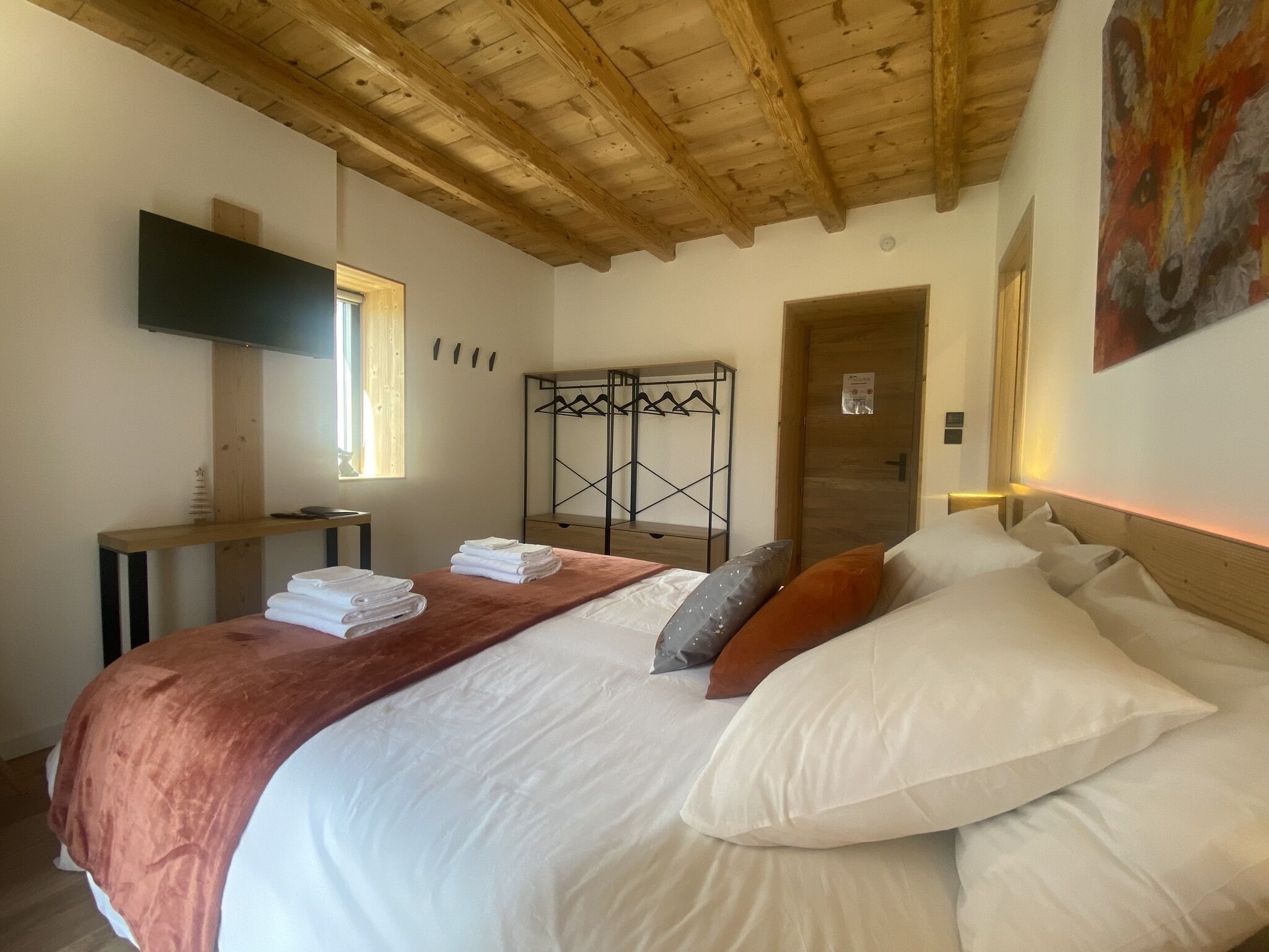 A bedroom with a wooden ceiling, a bed with white and brown bedding, a TV mounted on the wall, and a coat rack with hangers.