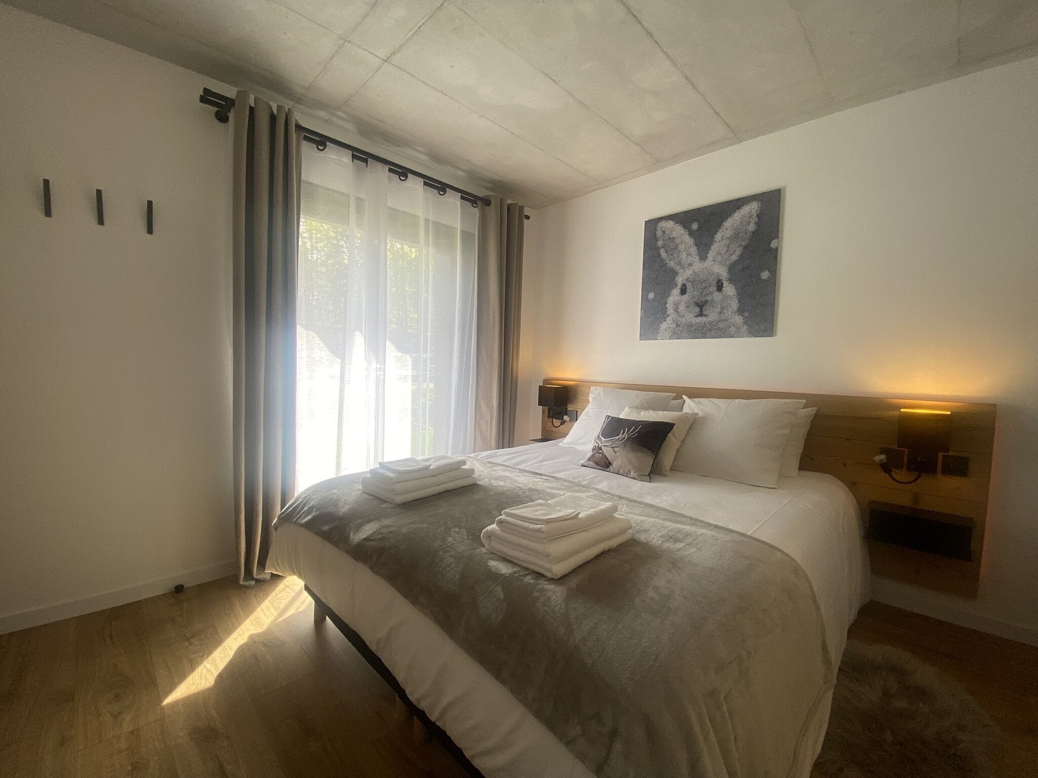 A neatly made bed with folded towels, a headboard with wall-mounted lights, and a wall art of a rabbit.