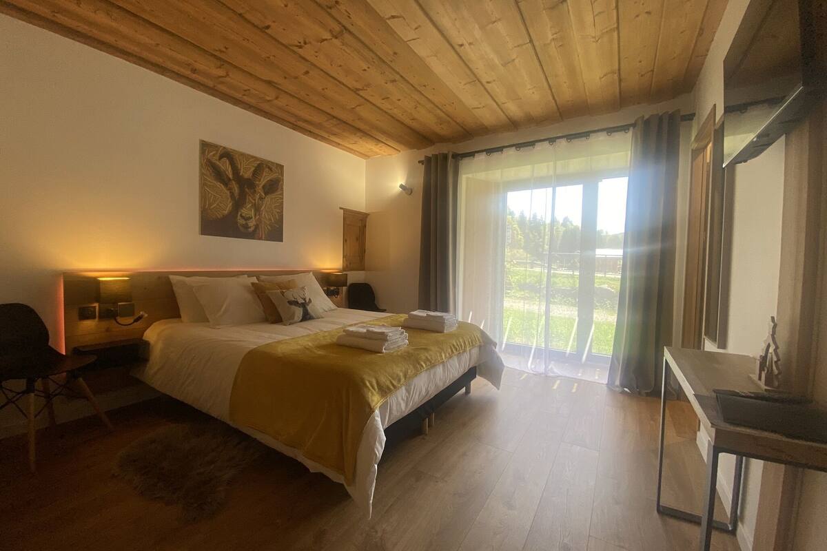 A bedroom with a wooden ceiling, a bed with a yellow bedspread, a nightstand with a lamp, a chair, and a view of a green landscape through a large window.